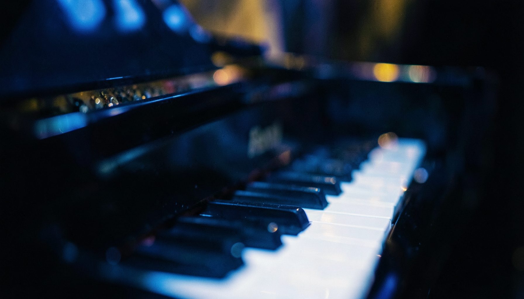 Close-up of a grand piano keyboard under soft blue and gold stage lighting with a shallow depth of field